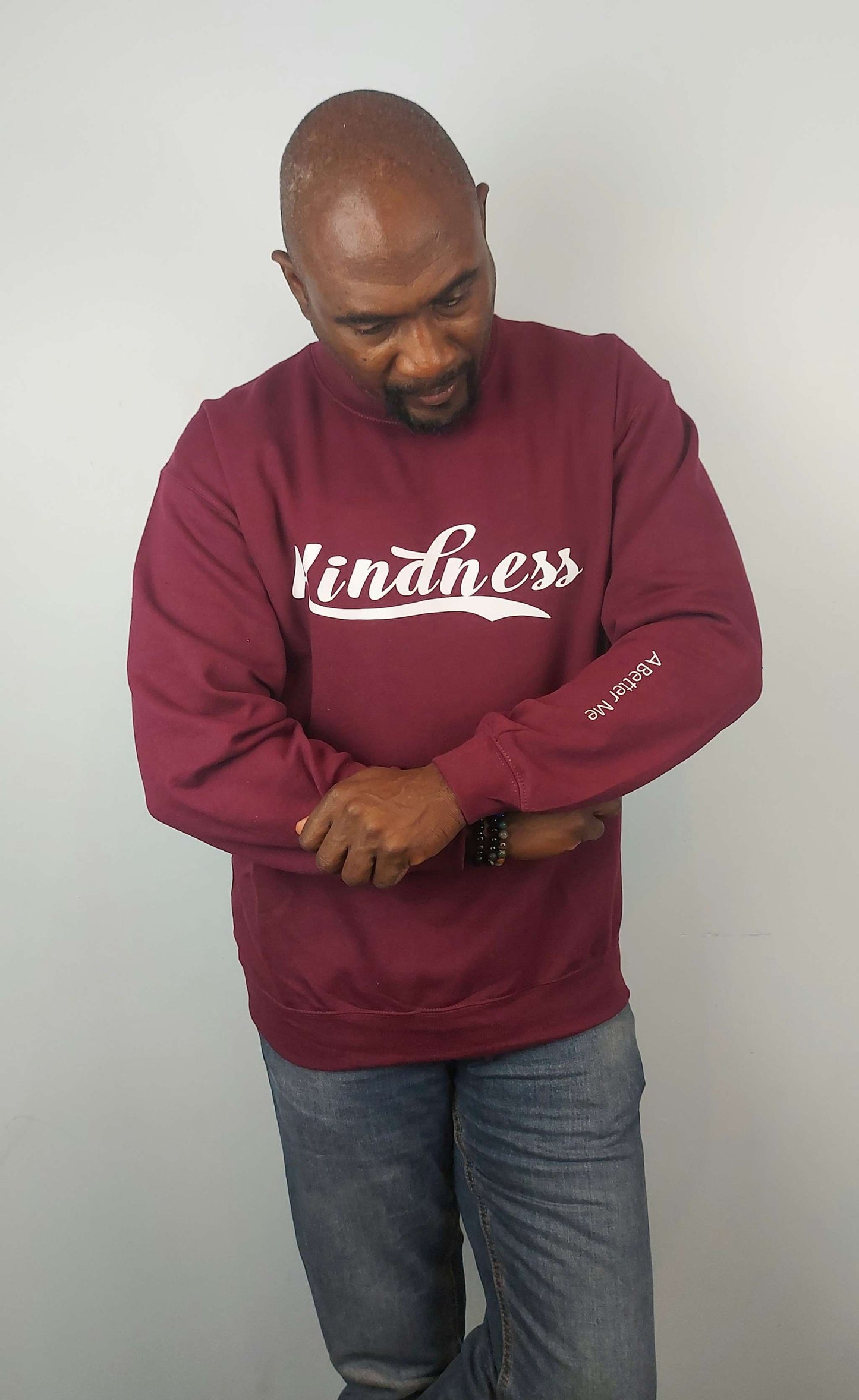 Burgundy crew neck sweatshirt with "Kindness" text in white, worn by a person with jeans.